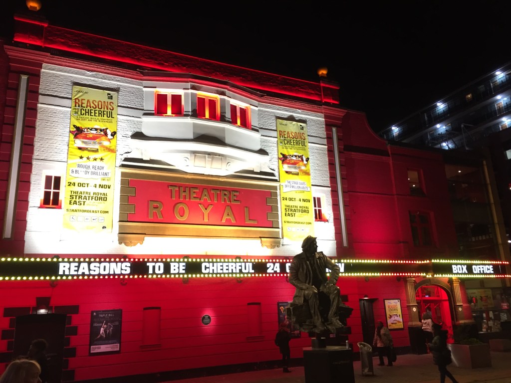 Exterior of Theatre Royal Stratford East, on which hang tall yellow banners advertising the Reasons To Be Cheerful musical. At the bottom, lit-up letters above the entrance doors show the musical name and running dates.