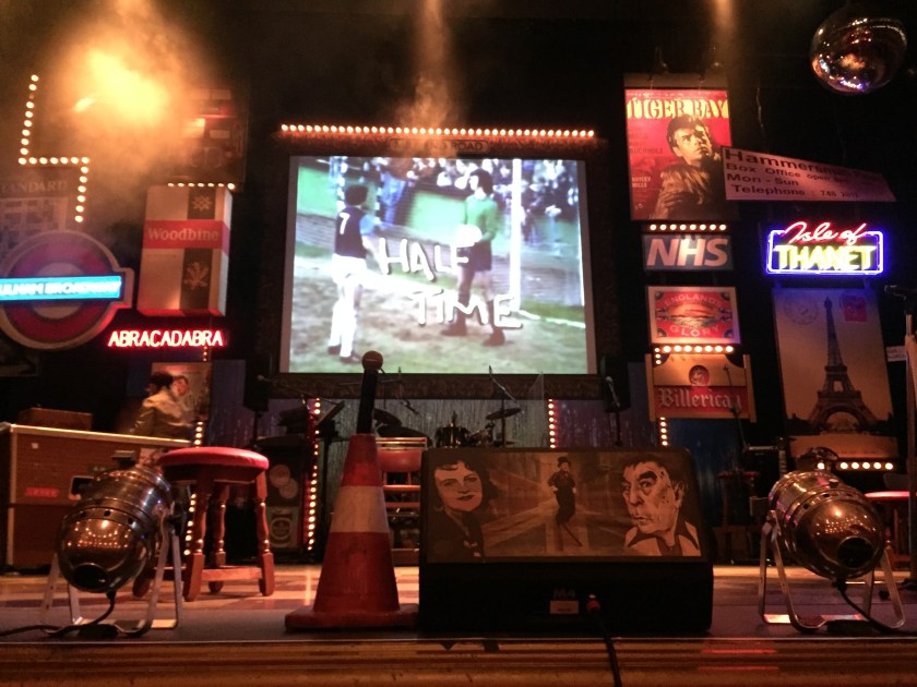 The stage during the interval. A screen at the back says Half Time over an image of footballers on a pitch. The stage has a few small neon-signs on the back, a pool table on the left, a drum kit at the centre back, and a traffic cone with a microphone on top at the front.