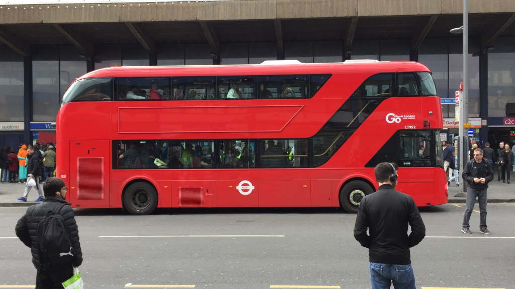 Red double-decker bus at a bus stop. The long bottom window turns upwards to follow the staircase up to the front of the top deck. So the long window on the left of the top-deck stops short before stairs. The window of the driver's cab is also separate to the other windows.