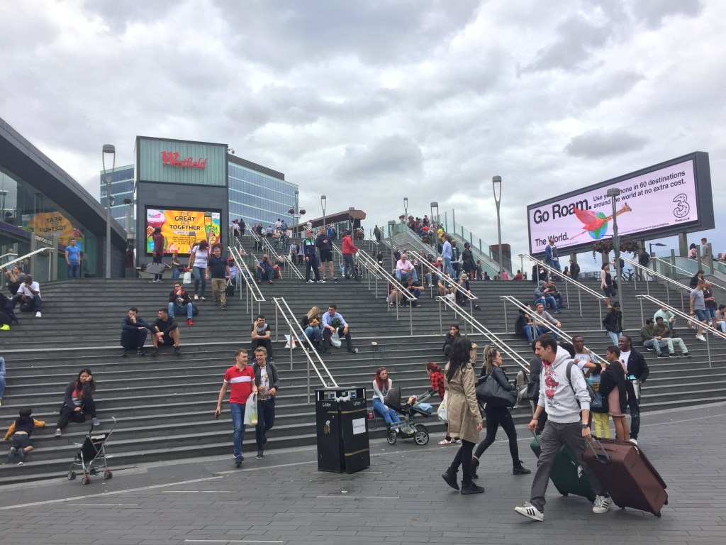 Very wide staircase outdoors, leading to Westfield Stratford shopping centre, with lots of people walking around or sitting on the steps.