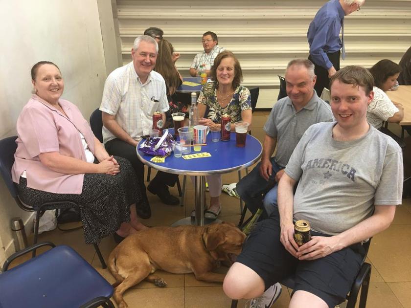 Me holding a can of drink while I sit with a group of people around a table, which has drinks and raffle tickets on it.