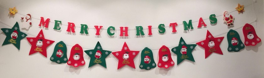 Festive banners hung on the wall on 2 lengths of string. The top string has alternating red and white letters spelling Merry Christmas. The lower string has alternating red and green stars and bells with Santa faces in their centres.