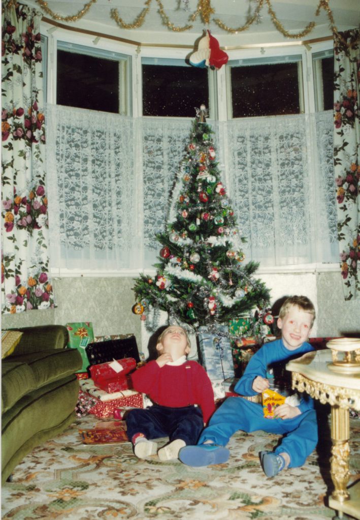 Me as a young child, sitting with my cousin next to a Christmas tree surrounded by presents in the lounge.