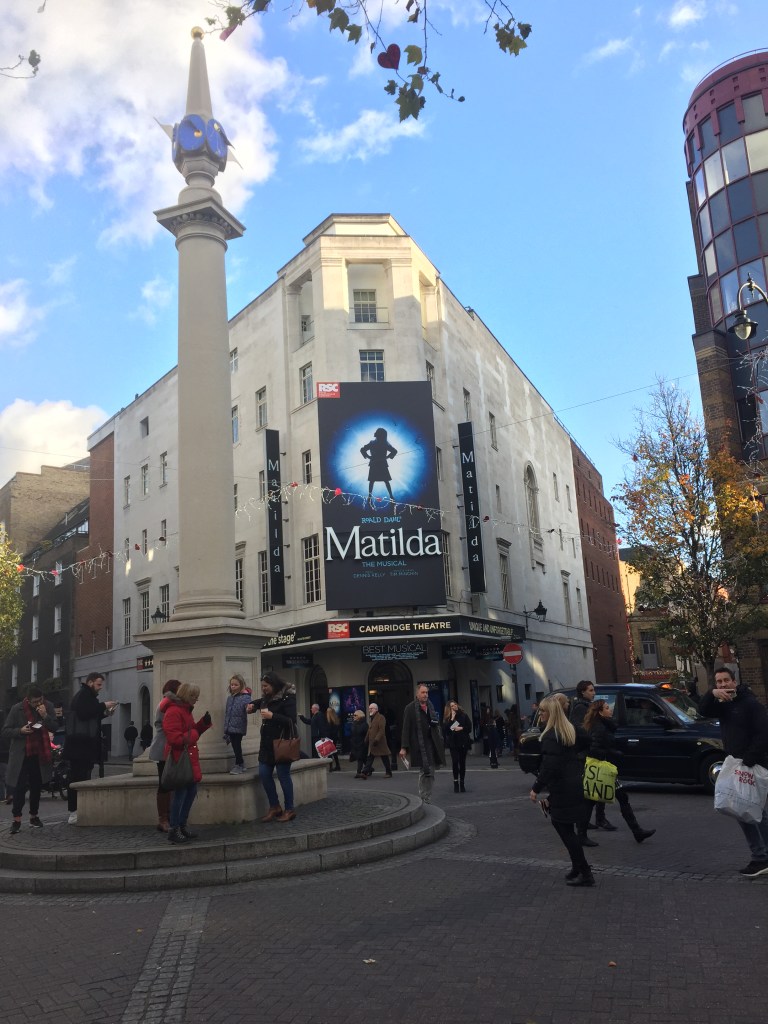 Seven Dials in London, a circular area with side streets in all directions. We face the Cambridge Theatre with a poster for Matilda The Musical over the entrance, featuring a silhouette of a defiant girl with her hands on her hips as she looks up to the side.