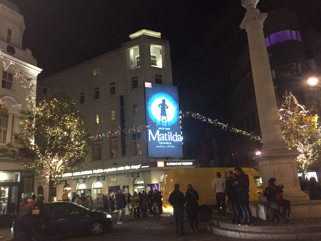 Seven Dials in London at night, a circular area with side streets in all directions. We face the Cambridge Theatre with a poster for Matilda The Musical over the entrance, featuring a silhouette of a defiant girl with her hands on her hips as she looks up to the side.