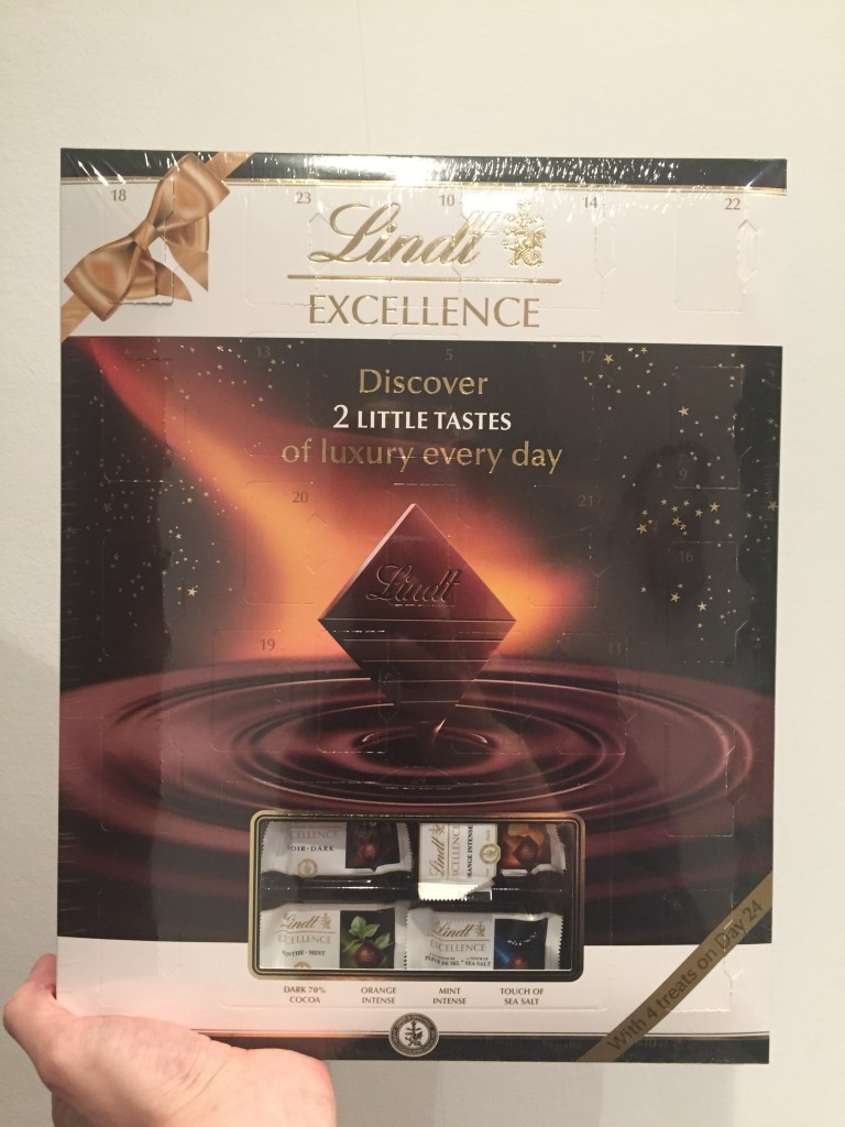 Lindt Excellence Advent Calendar - Discover 2 little tastes of luxury every day. The large background of the calendar shows a square piece of chocolate being dipped into a rippling pool of chocolate.
