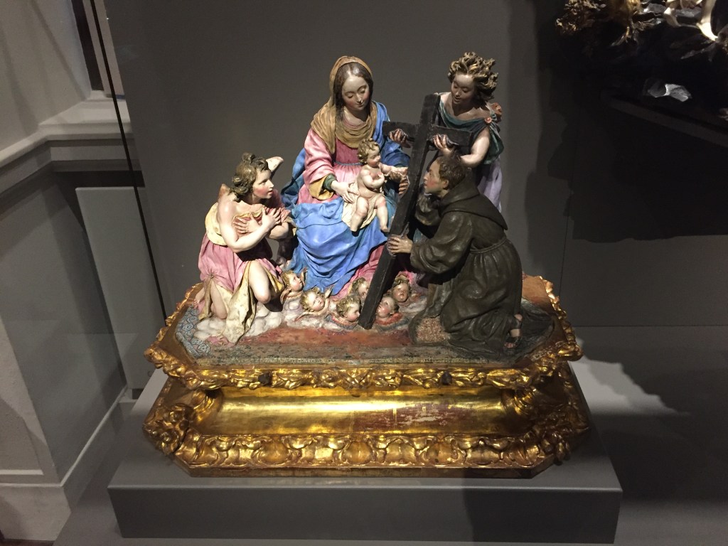 A colour model, on an ornate gold base, showing the Virgin Mary in a blue robe, with the baby Jesus on her lap. On the right, a lady hold the top part of a long cross, while a kneeling man holds the lower end. To the left, a young boy sits with his arms crossed over his chest.