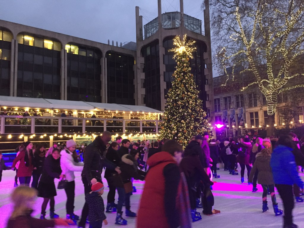 On the ice rink in front of the Natural History Museum, people skate around a tall Christmas tree, lit by thousands of small lights, with a bright star on the top. A tree to the right, outside the rink, that lost its leaves in the autumn, has lights all over its trunk and branches.