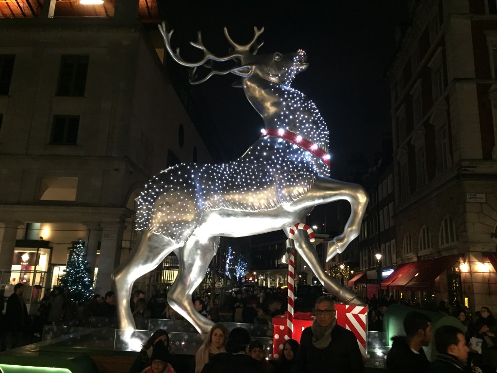 A shiny metal sculpture of a reindeer covered in tiny lights. It's lifting its right leg and looking upwards as if about to take off and fly.