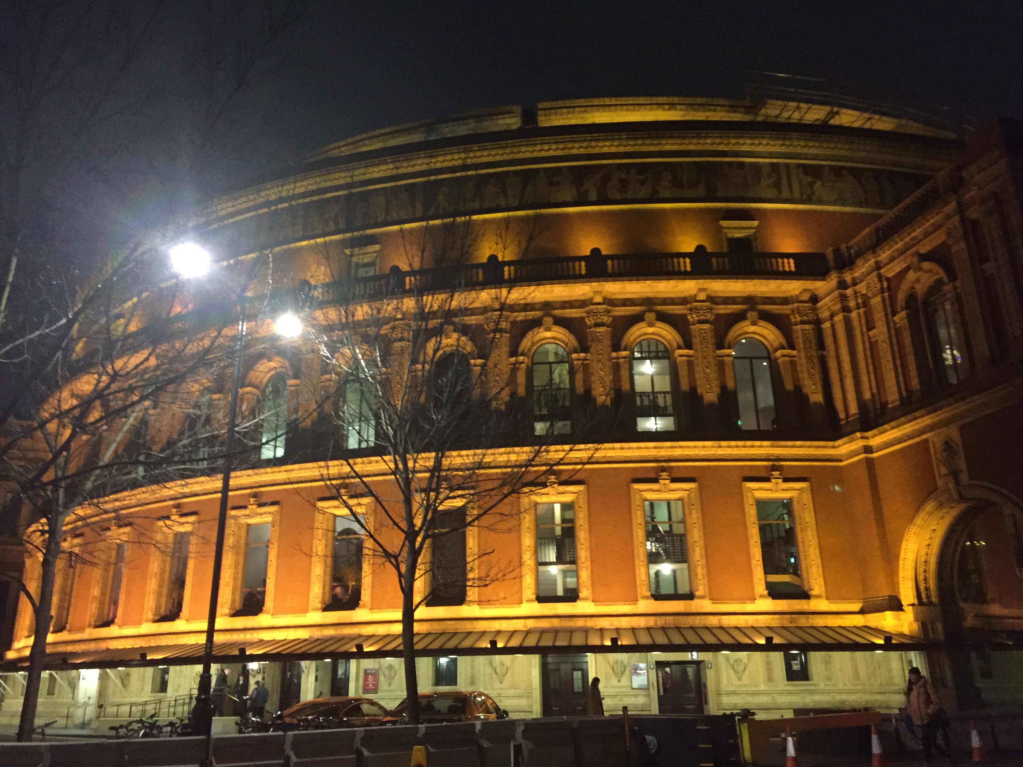 Outside view of the Royal Albert Hall, a huge circular building, appearing golden in colour as it's lit up in the night sky.