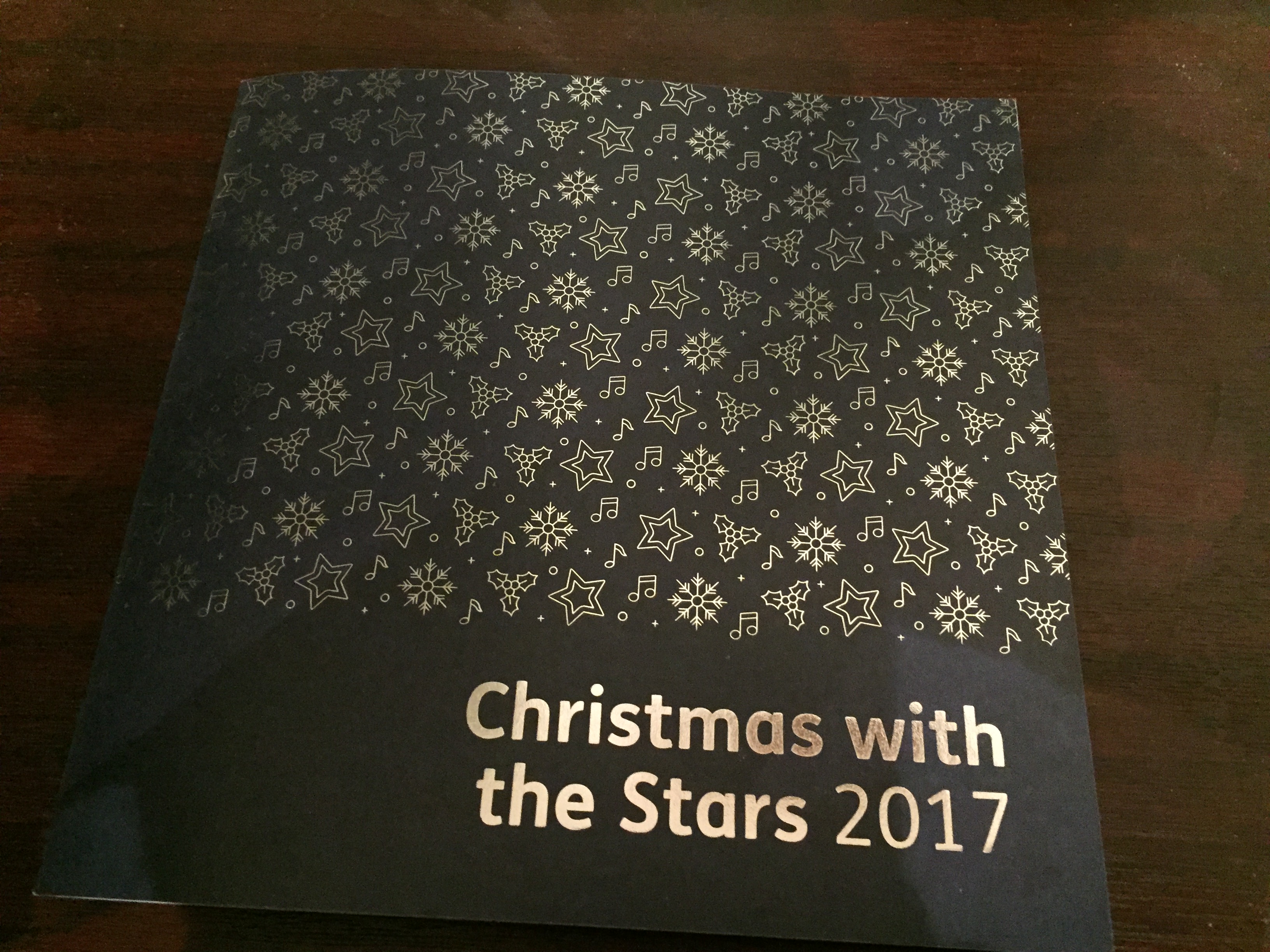 Programme for Christmas With The Stars 2017. The cover is navy, with the event title in silver letters in the bottom right. The rest of the cover above it is filled with a mixture of silver stars, holly, snowflakes and musical notes.