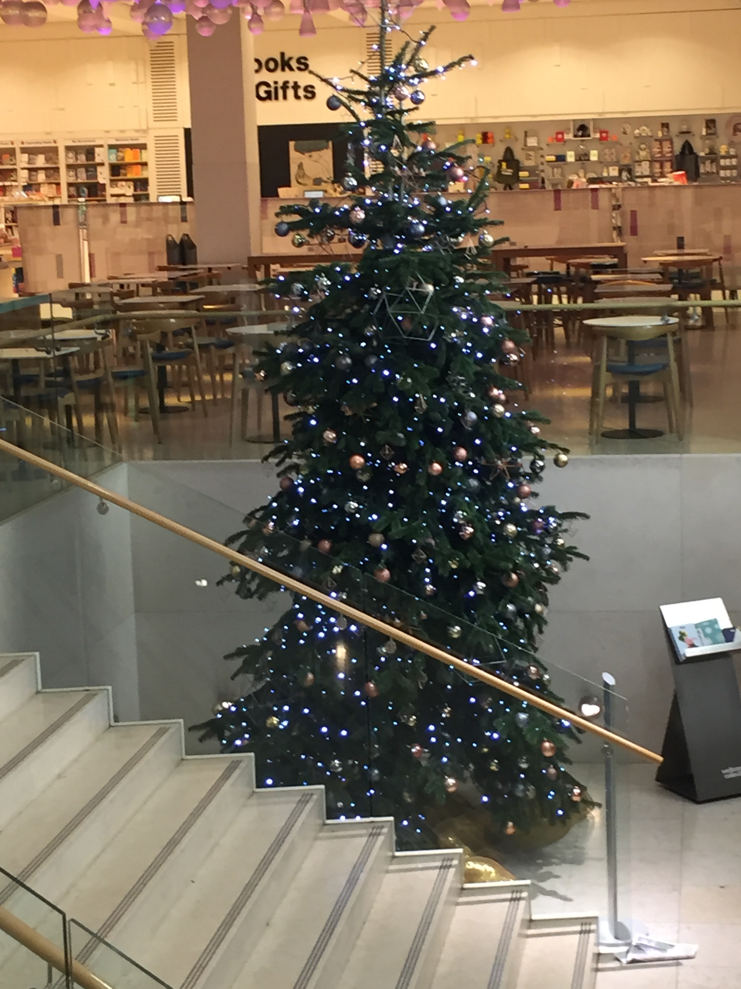 Tall green Christmas tree covered in hundreds of small baubles of various colours, situated beside a staircase.