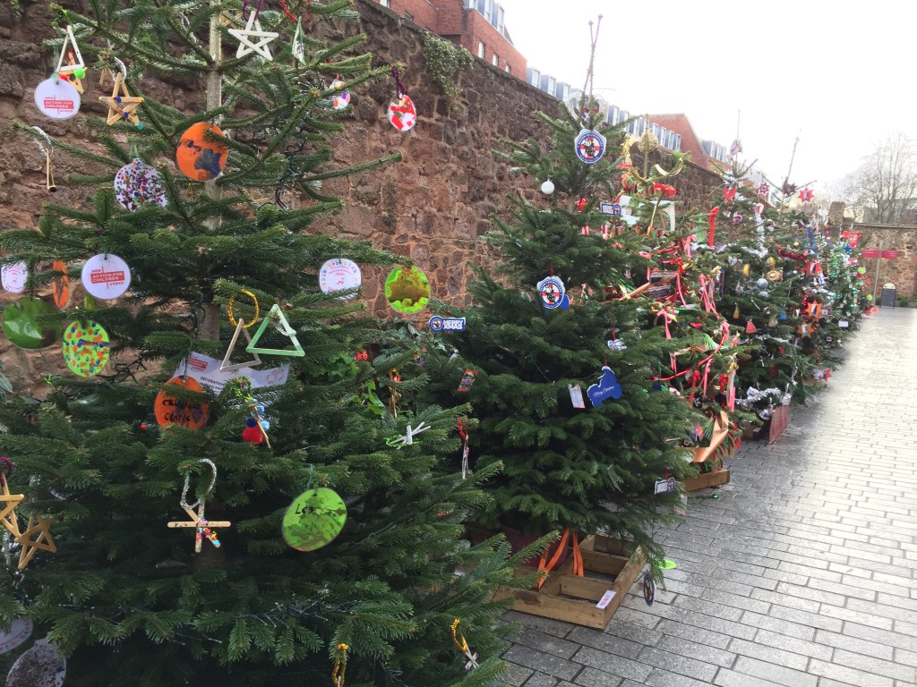 A long line of decorated Christmas trees along a wall by the pavement, each with labels representing a different charity.
