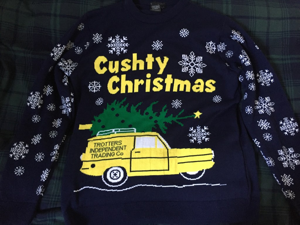 Navy jumper covered in a variety of patterned snowflakes. At the top of the chest is Cushty Christmas written in big yellow letters. Below this is the Trotters yellow three-wheeled Reliant Regal van, with a green Christmas tree on the roof rack.