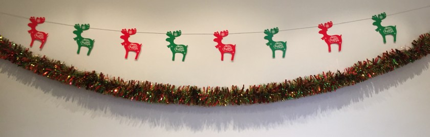 Threaded along a line of string on the wall are alternating red and green cutouts of reindeer with Christmas greetings written on them. Below them is a long length of golden brown tinsel along the wall.