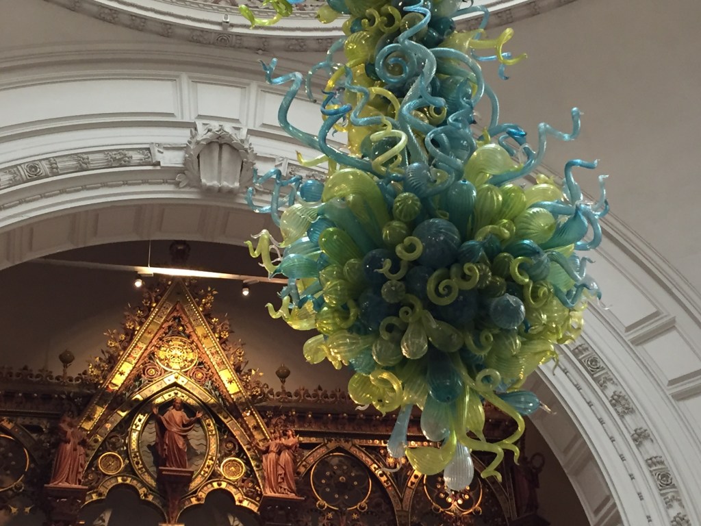 Chandelier in the main hall of the V&A Museum. It has many curved intertwining tubes with bulbous ends, in a mixture of blue and green colours, forming a teardrop-like shape overall.
