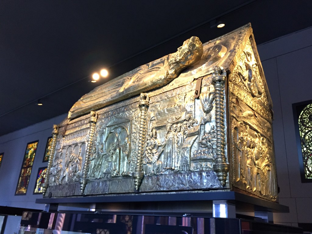 A massive and very ornate gold structure in the shape of long building rectangular building with a sloping roof. It's covered in embossed scenes depicting a variety of people and objects.