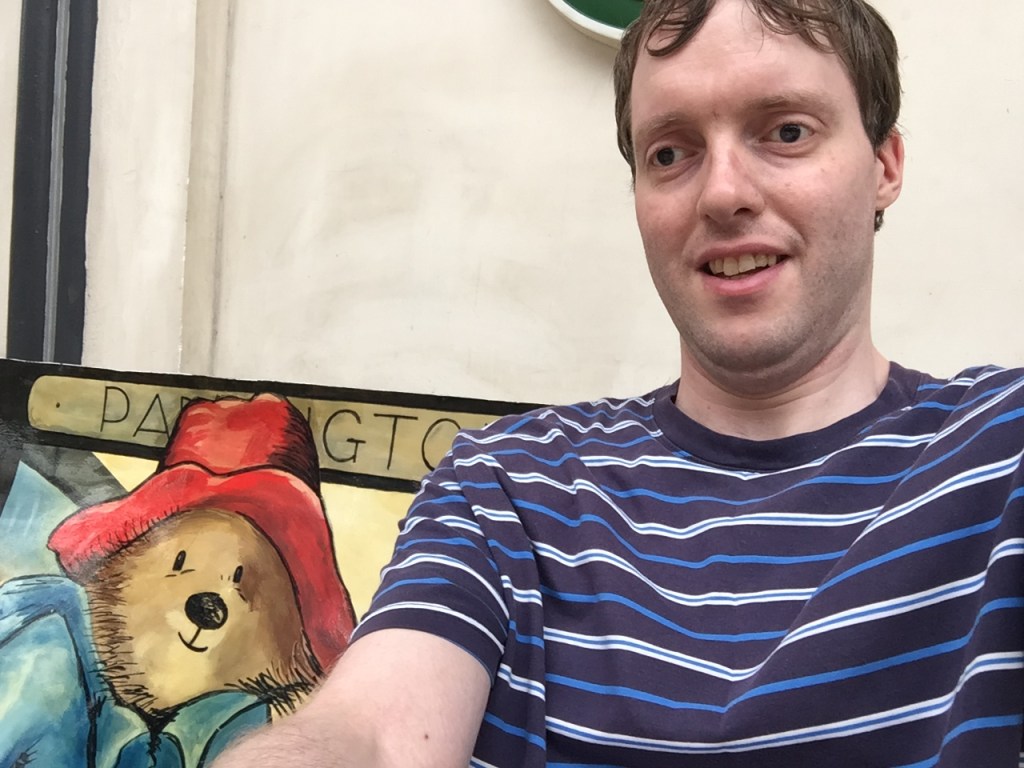 Glen sitting on a bench that's decorated with a painting of Paddington Bear, so it looks like they're sitting next to each other and both smiling at the camera.