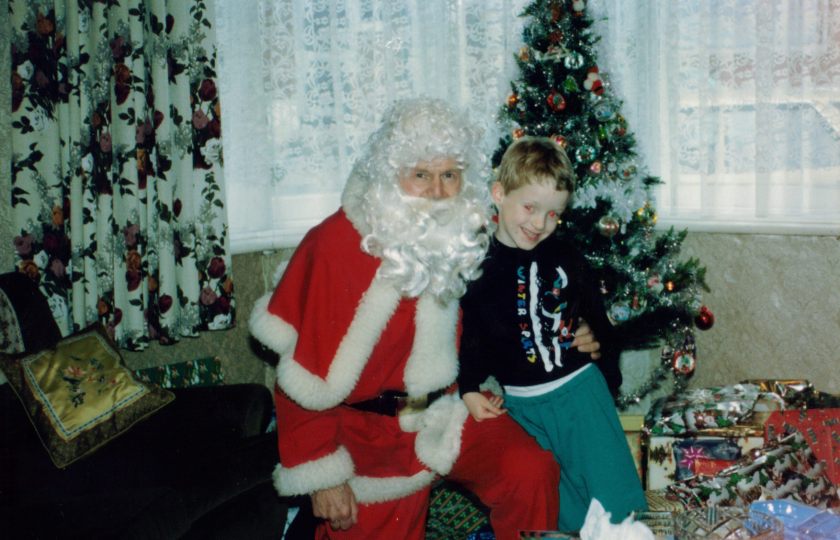 Me as a young child, smiling and leaning against Santa Claus, who has his arm around me. It's my grandad dressed up. We're in front of the Christmas tree and presents, next to the living room window.