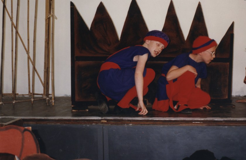 Me and another boy, dressed in blue robes and red leggings, with blue and red hats, crouching on stage next to a spiky wall, hiding from the people we're spying on.