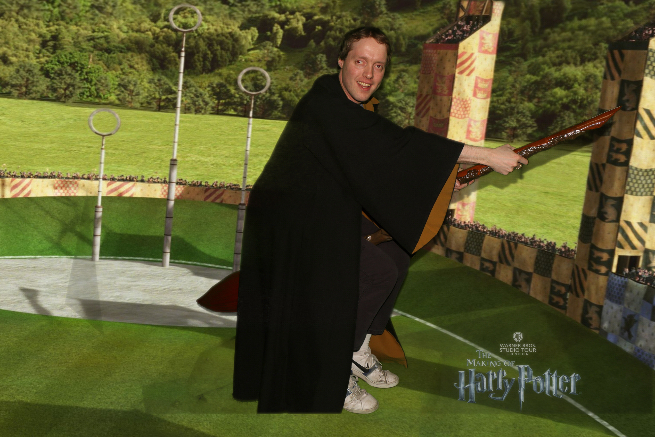 Side view of Glen smiling in a brown robe, as he flies on a broomstick past the Quidditch Arena at Hogwarts, which has hoops on very tall poles, and lots of grass and trees in the background.