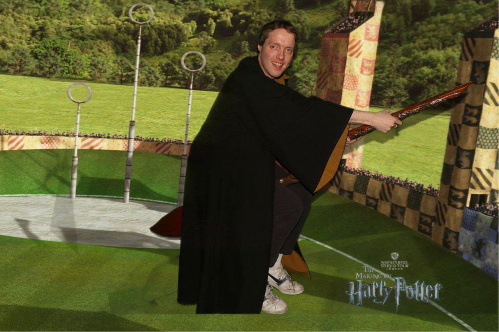 Side view of Glen smiling in a brown robe, as he flies on a broomstick past the Quidditch Arena at Hogwarts, which has hoops on very tall poles, and lots of grass and trees in the background.