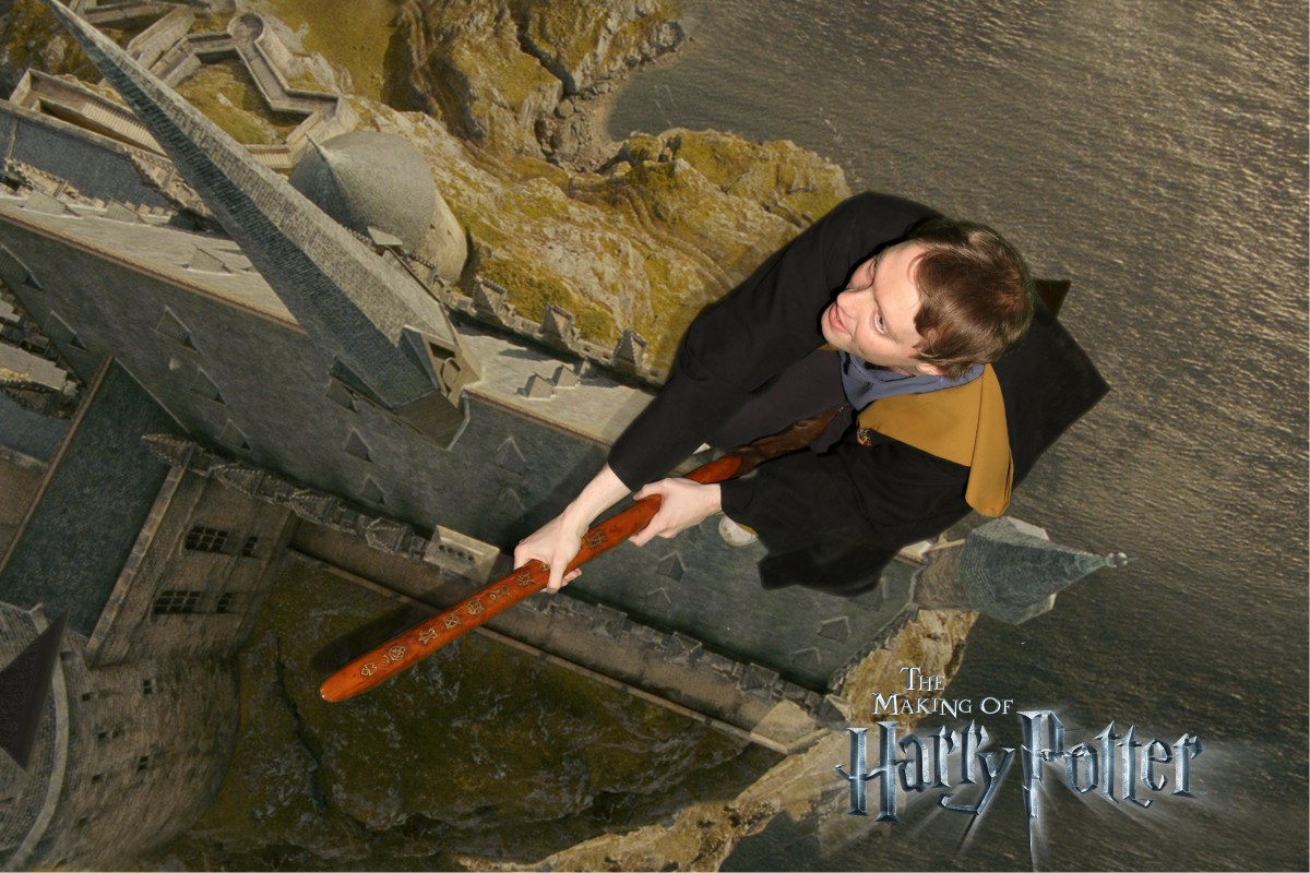 Overhead view of Glen in a brown robe with yellow inner lining, flying on a broomstick over Hogwarts Castle.