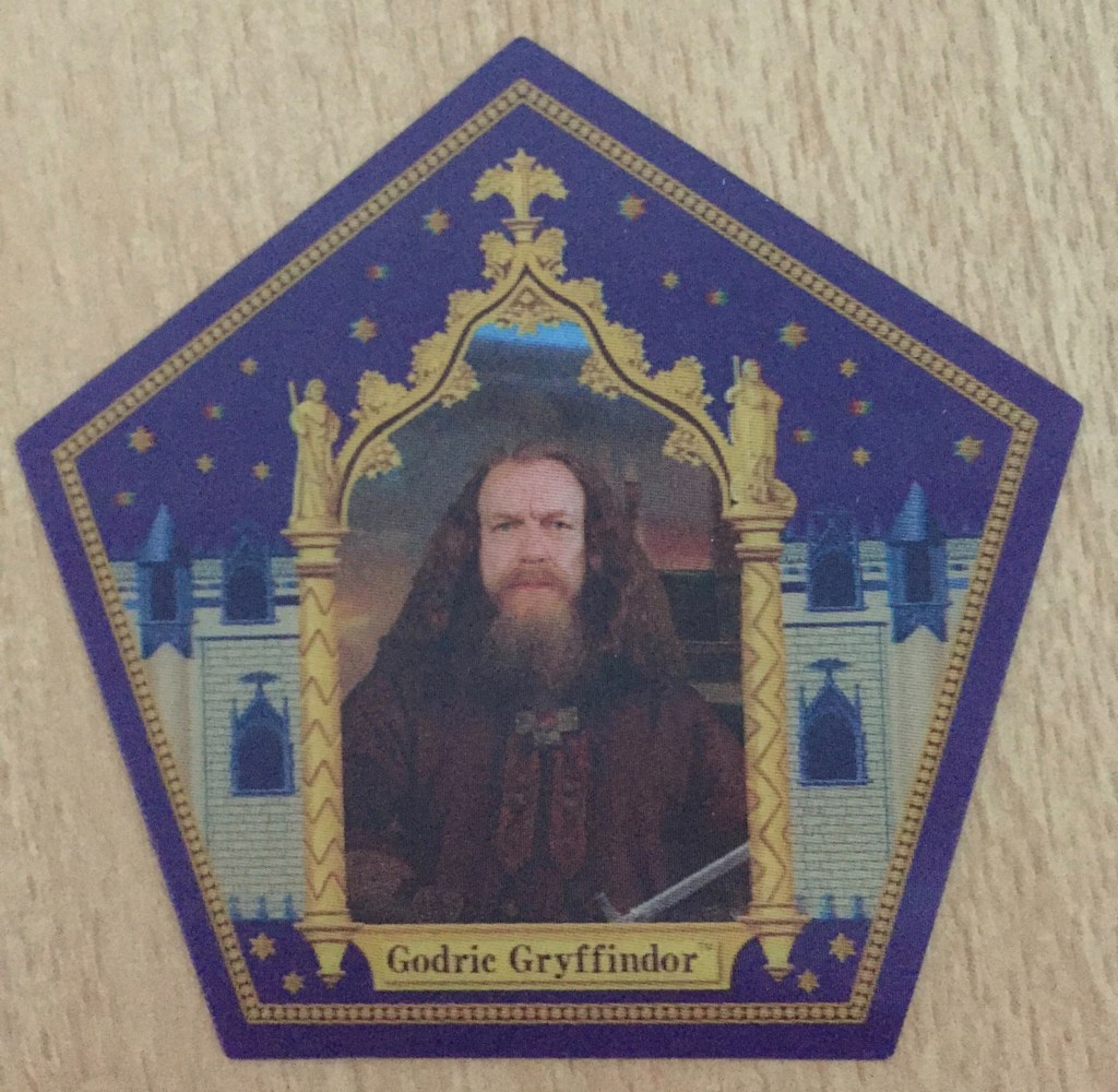 5-sided card with a blue background and gold detailing, featuring the bearded Godric Gryffindor in an ornate gold frame in the centre, part of his sword just visible at the bottom of the frame.