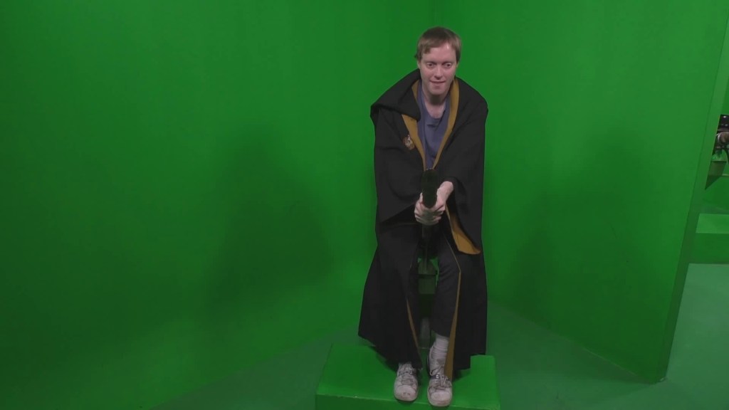 In front of a plain green background, Glen is wearing a brown robe with a yellow inner lining visible, while mounted on a broomstick as if ready to fly, with his hands clasped around the handle at the front.