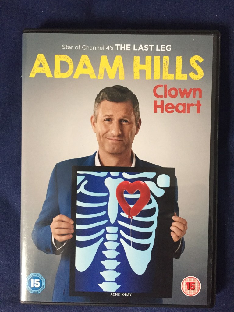 DVD case for Adam Hills - Clown Heart. Shows Adam smiling while holding up a mock X-ray in front of his chest. The X-ray shows a spine and ribcage, with a drawing of a long red balloon tied into a heart shape, floating in front of which his regular heart would be.