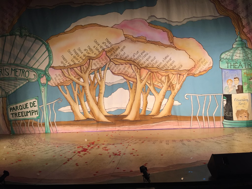 Stage backdrop artwork, a group of large cream coloured trees, with the text Once Upon A Time and Happy Ever After on some of their leafed tops. To their left are signs for the Parque De Treeumph and Paris Metro. And to their right is a sign saying Theatre.