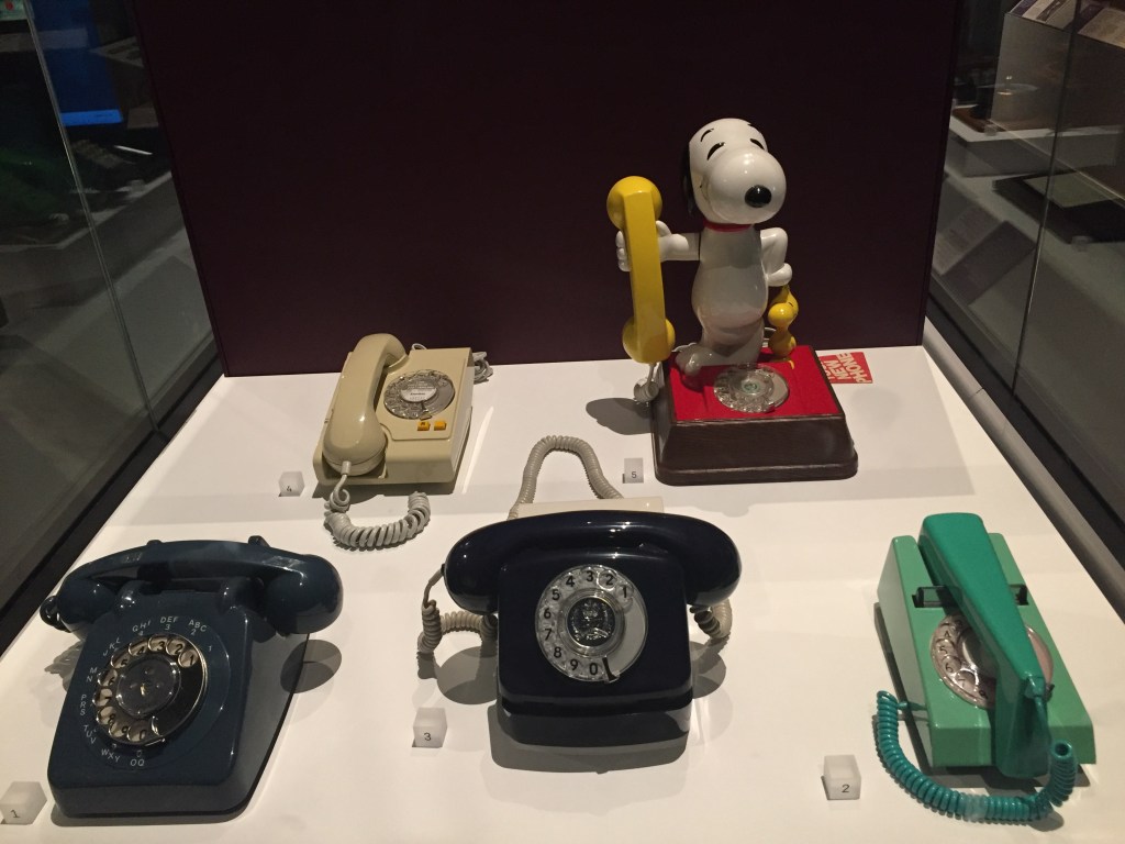 A varied selection of 5 old style telephones with rotary dials. The most distinctive is one that has a model of Snoopy the dog standing up on the red base that has the dial, and holding the yellow receiver in his right hand.