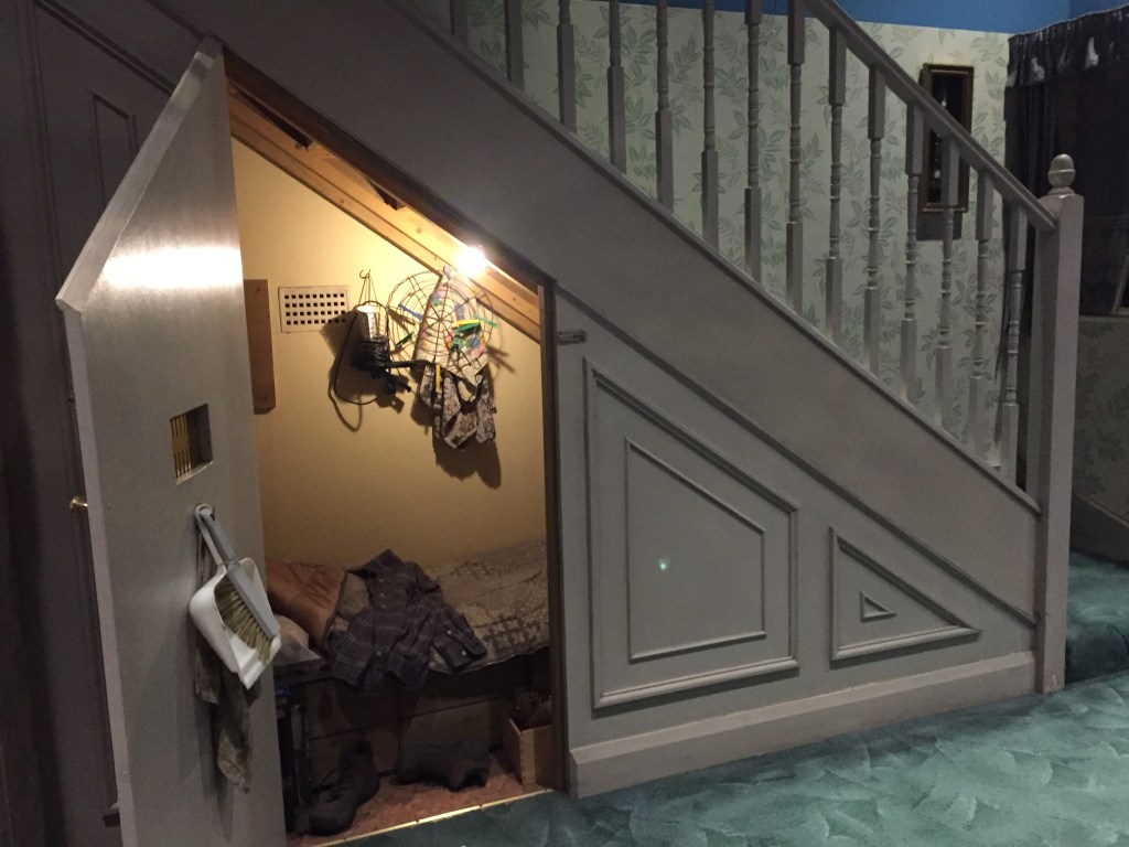 An open cupboard under a while staircase, with the inner light switched on to see the contents, including a small bed and items of clothing, and a dustpan and brush hanging on the inside of the door.