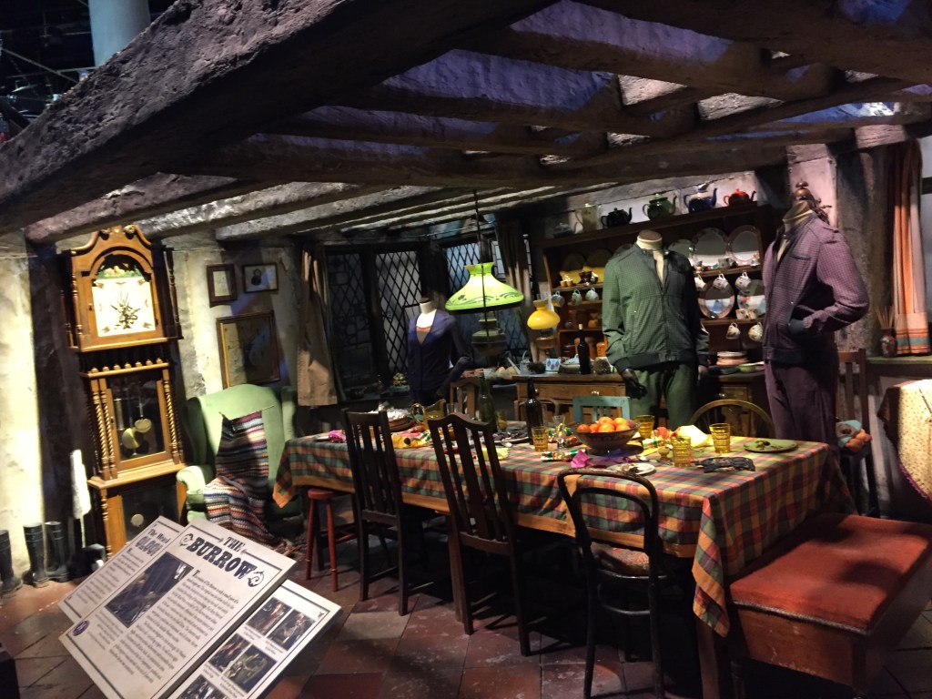 Inside The Burrow, home of the Weasleys, including wooden beams on the ceiling, a long dining table, headless mannequins wearing character's costumes, and a tall ornate clock with a pendulum.
