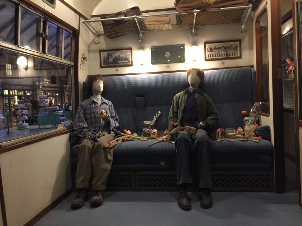 Models of Harry Potter and Ron Weasley sitting inside a carriage on the Hogwarts Express, each holding a box of Bertie Botts Every Flavour Beans, with a messy pile of other sweets on the seat between them.