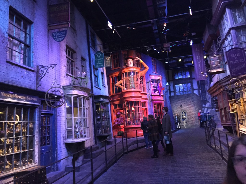The set of Diagon Alley, with various shops on each side of the narrow cobbled street. One shop has a huge model of man standing on the ground floor, with his head sticking out of the roof above the first floor. His arms come out of the sides of the first floor, one of which holding the brim of the top hat on his head, as if he's about to doff his hat to passers by.