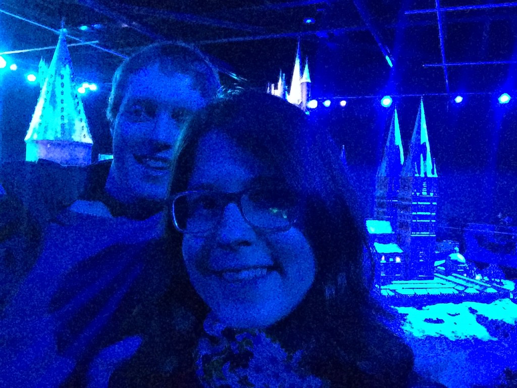 Glen and a lady wearing glasses posing for a selfie in front of the large Hogwarts castle model, which is dusted with snow. The entire scene is bathed in blue light.