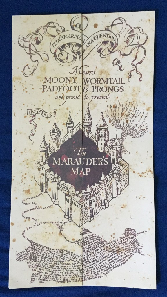 The folded-up Marauder's Map, tall and cream coloured, with decorative artwork in black ink. Near the top is the text Messrs Moony, Wormtail, Padfoot and Prongs are proud to present. Below this is a 4 walled square castle with the text The Marauder's Map in the centre on a black background.