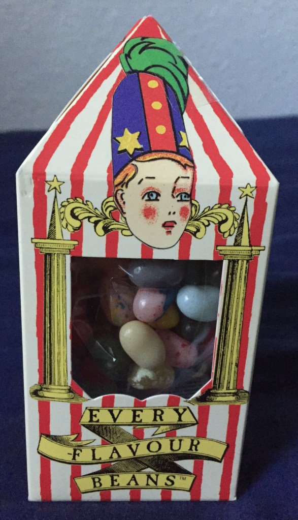 Red and white striped pack for Bertie Botts Every Flavour Beans, looking a bit like a tall, narrow circus tent. The rosy cheeked face of Bertie, wearing a tall blue and red hat, sits above the plastic window showing various different coloured beans.