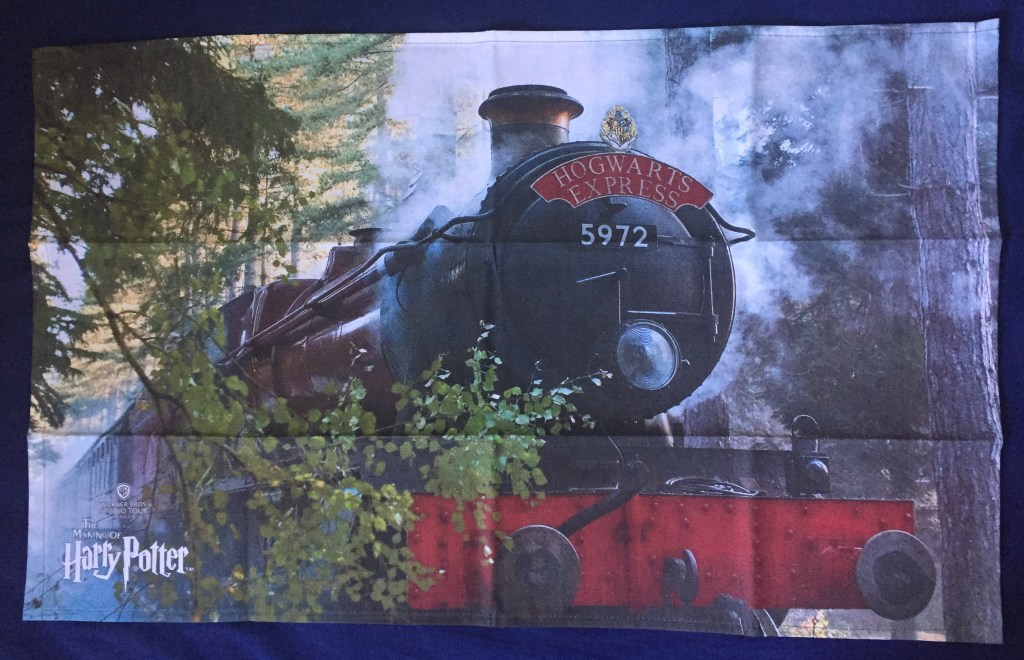 Tea towel showing a large image of the Hogwarts Express steam train passing by trees that line the edge of the railway.