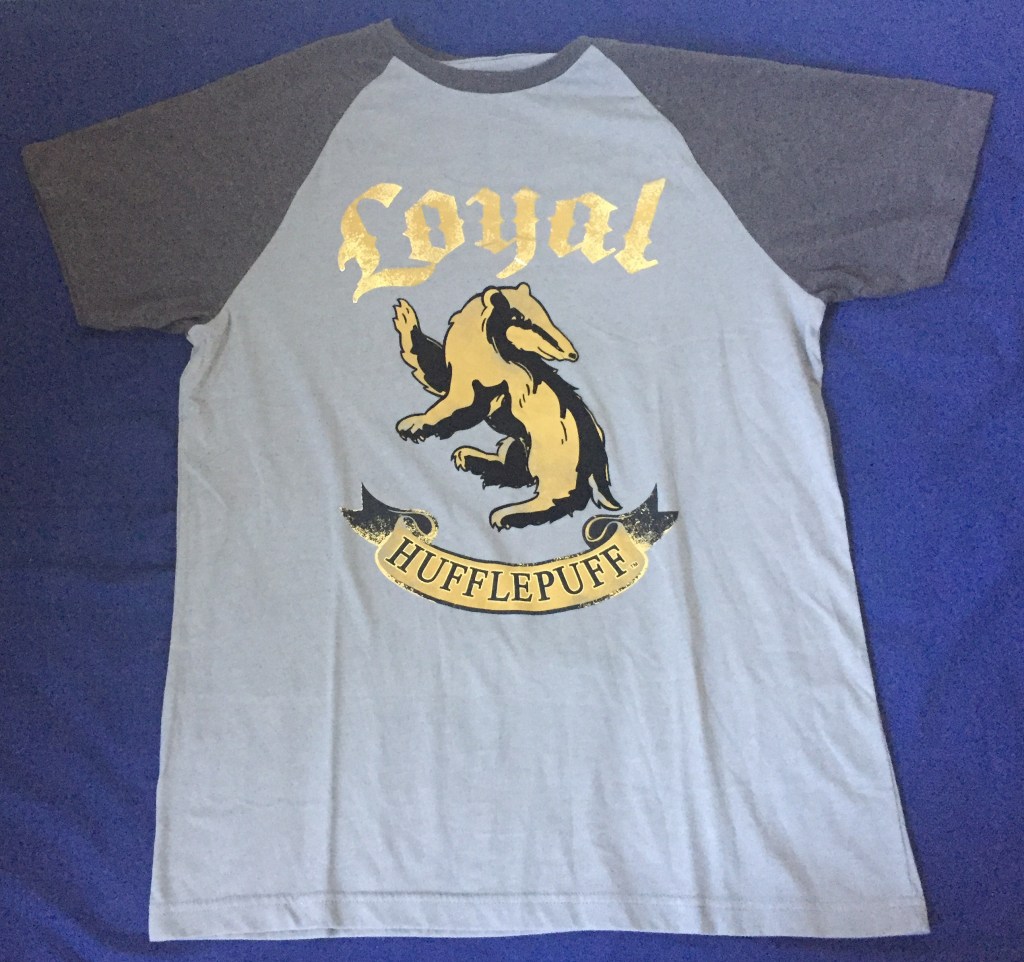 T-shirt with light grey body and dark grey short sleeves. The body has the word Loyal at the top in gold letters, below which is a large fold and black badger, and below that is a scroll across the chest with the word Hufflepuff.