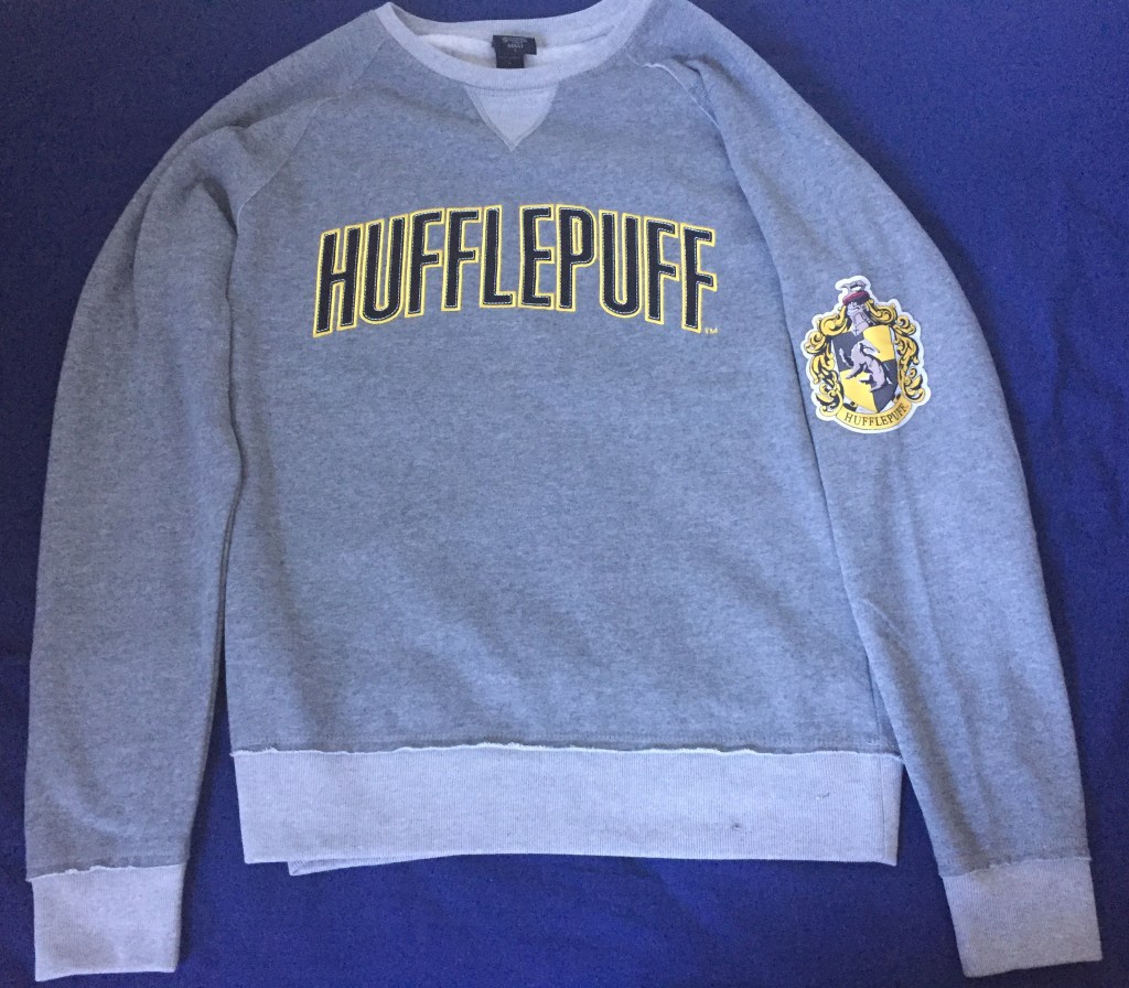 Grey sweater with the word Hufflepuff across the chest, in large black letters with gold edging. The official badge of Hufflepuff is on one sleeve, featuring a badger in the centre.