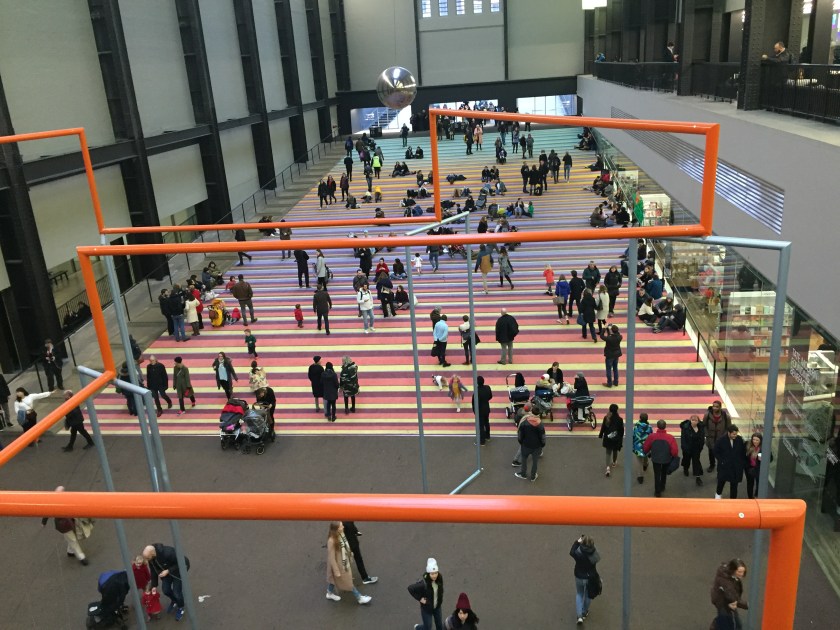 The long, wide entrance slope of the Turbine Hall at the Tate Modern, adorned with a multi-colour striped carpet on which people are sitting as others walk past. Above them, a large metal ball swings like a pendulum.
