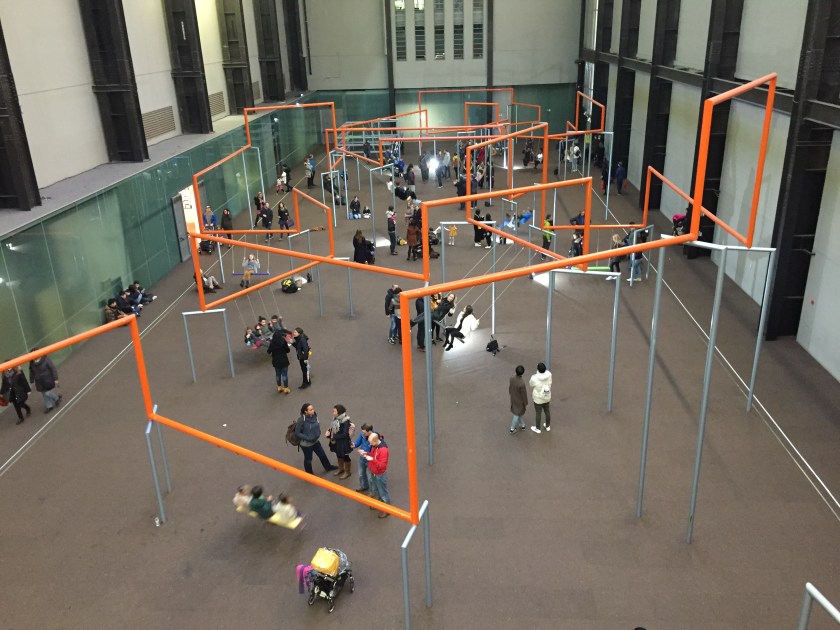 In the Turbine Hall of the Tate Modern, people are sitting on swings hanging down from orange bars, which criss-cross the space in a seemingly random pattern, with the seat of each swing holding 3 people.