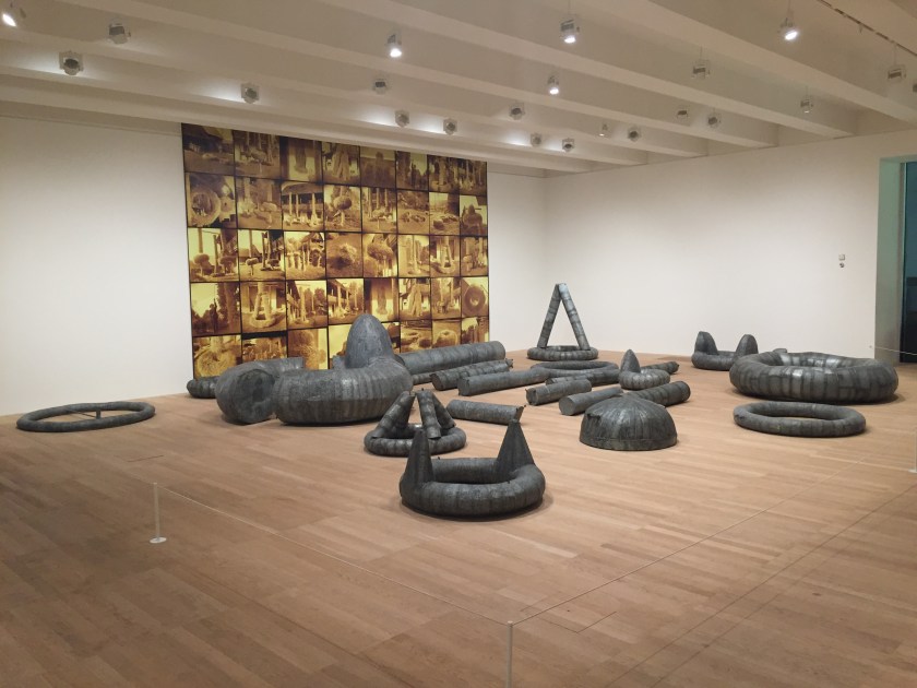 A mixture of large, cylindrical, grey metal cases, some straight cylinders and some circular ones, in front of a wall-sized grid of 40 sepia photo, documenting the making of the items originally inside the casings.