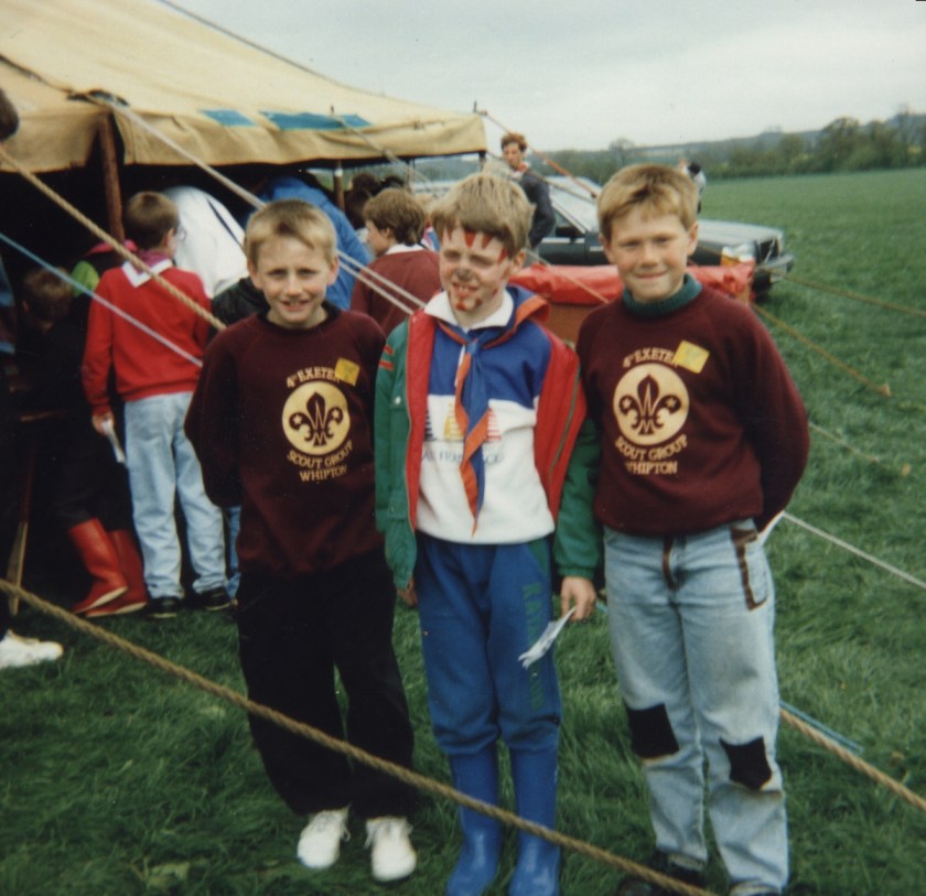 Me in the scouts as a kid, wearing blue trousers, a top that's half blue and half white, a green coat, and a blue and orange neckerchief. My face is painted with triangular marks all along my jaw (pointing up) and forehead (pointing down), and my nose is also painted. I'm standing in a field with 2 boys, who are wearing dark red sweaters with the Scout Association logo on.