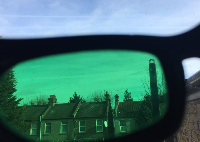 The blue sky and a row of houses below, as viewed through a green lens of my sunglasses. The glasses lens takes up most of the image, but above the glasses at the top is a small section of the blue sky as it normally looks, for comparison.