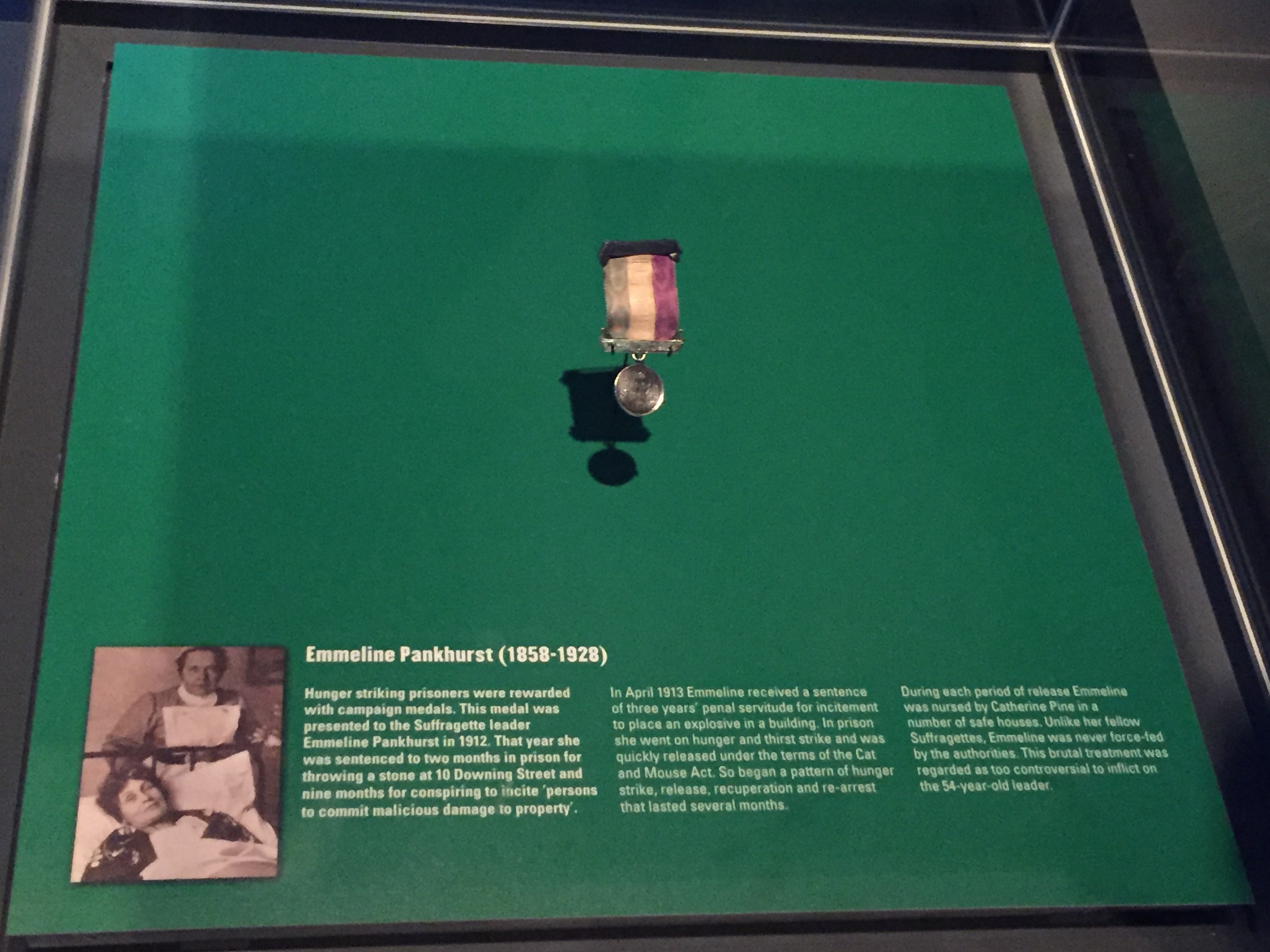 Medal awarded to Suffragette leader Emmeline Pankhurst. The small round silver medal has a small ribbon attached to the top with 3 vertical stripes in what appears to be green, yellow and red.