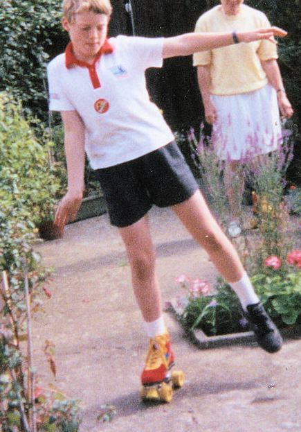 Me as a child in our back garden, wearing a white t-shirt and black shorts, reaching my arm out to the side as I balance on a red and yellow roller skate on my right foot. I'm wearing a regular black shoe on my left foot. My mother is watching in the background.