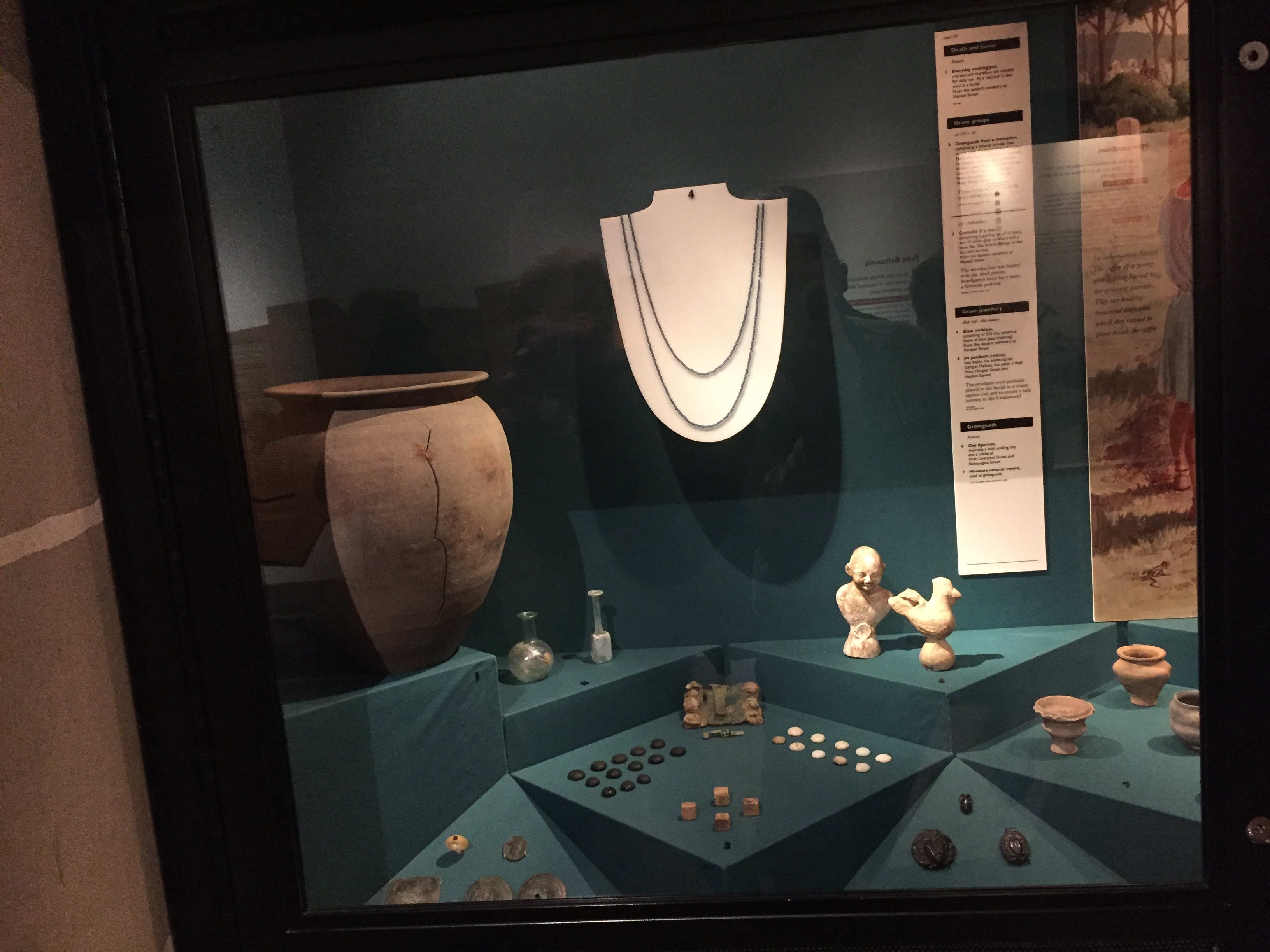 Display case with objects including a necklace, a large pot, coins, and other small artefacts. The wall and floor of the case are a dark blue colour, and each object has a small black number near it. A tall white panel at the back of the case has very small text describing each object.